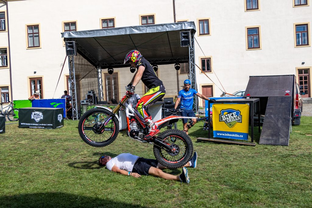 Motocyklový kaskadér přejíždí na zadním kole nad dobrovolníkem ležícím na trávě.