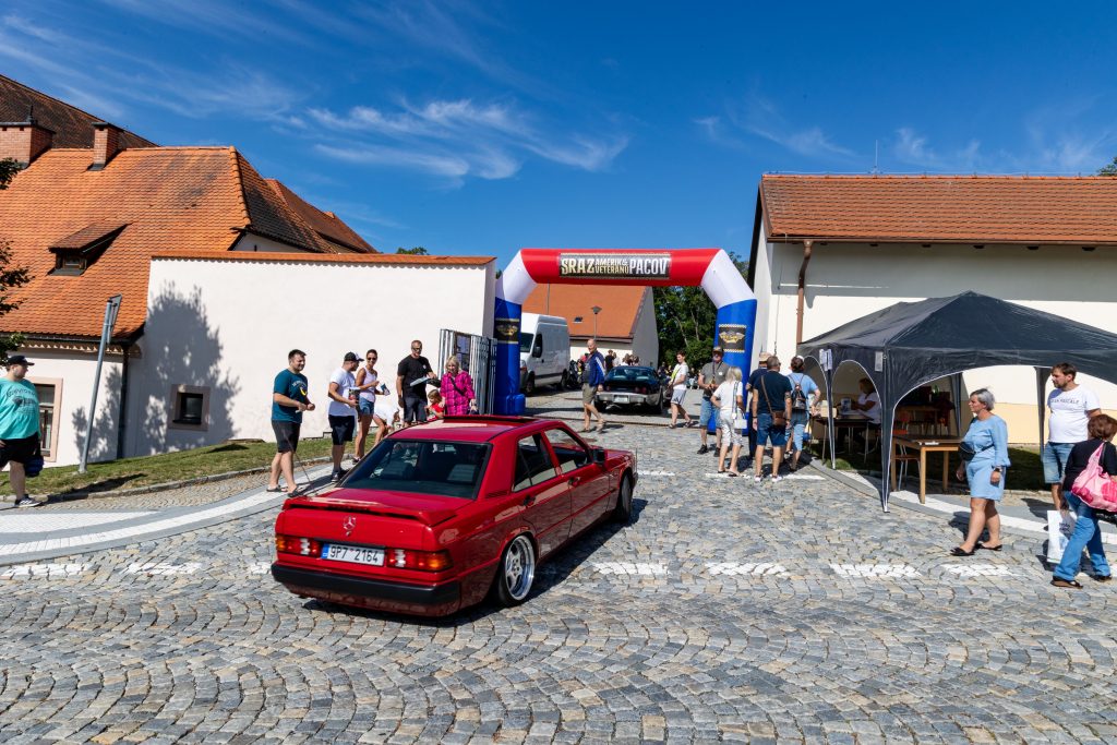 Červený upravený vůz projíždí pod nafukovací startovní bránou mezi diváky.
