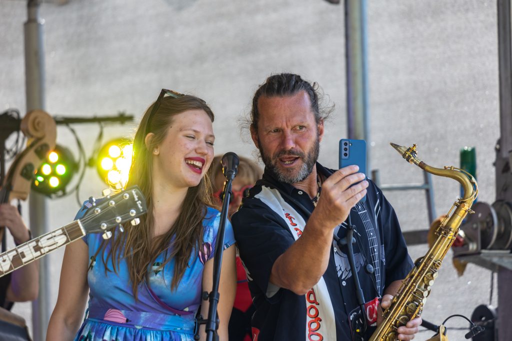 Mladá zpěvačka se směje u mikrofonu vedle saxofonisty, který v ruce drží mobilní telefon, na denním pódiu.