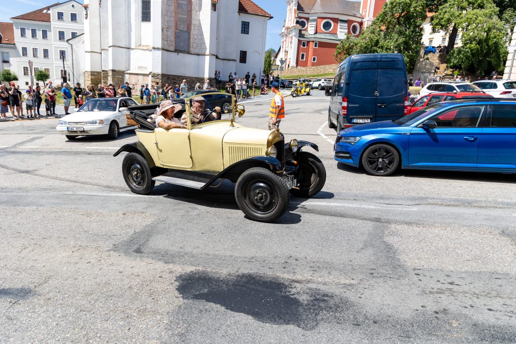 Posádka béžového historického automobilu projíždí městskou ulicí mezi diváky.