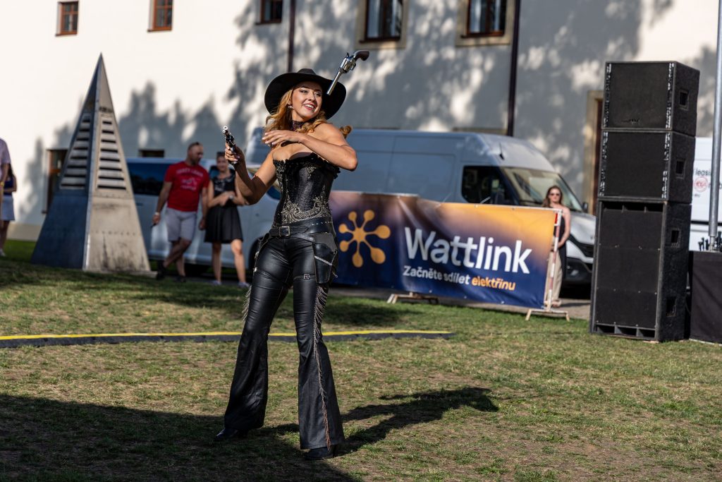 Performerka v černém westernovém kostýmu a klobouku drží v každé ruce zdvižený revolver.