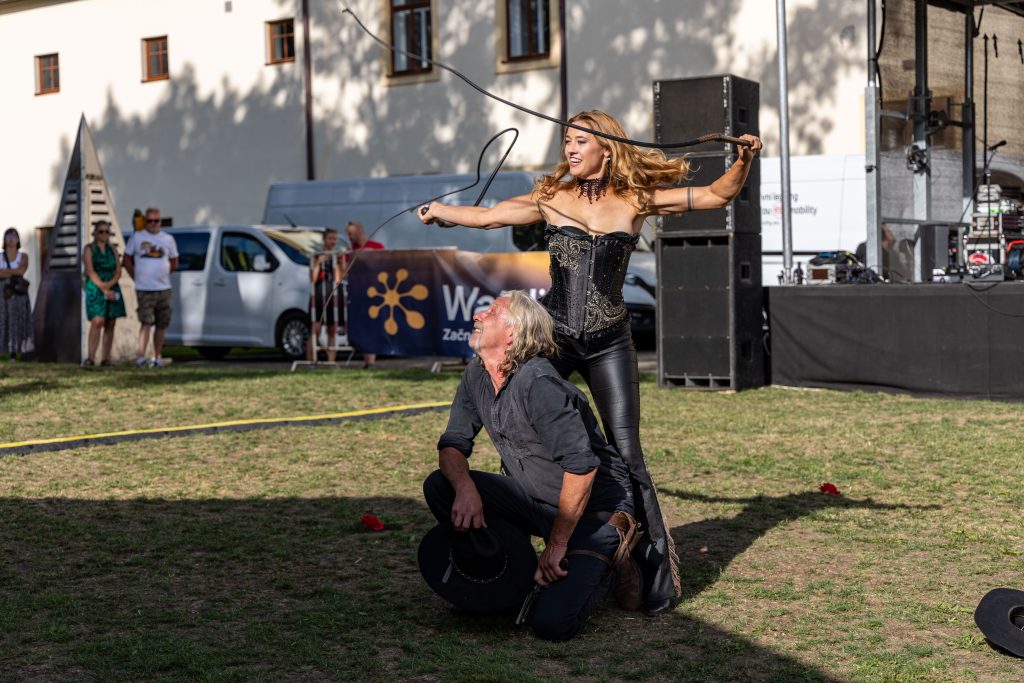 Performerka v černém westernovém kostýmu švihá bičem nad klečícím mužem v černém klobouku.