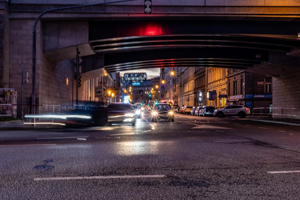 Noční městská ulice pod železničním mostem se světelnými stopami projíždějících aut.