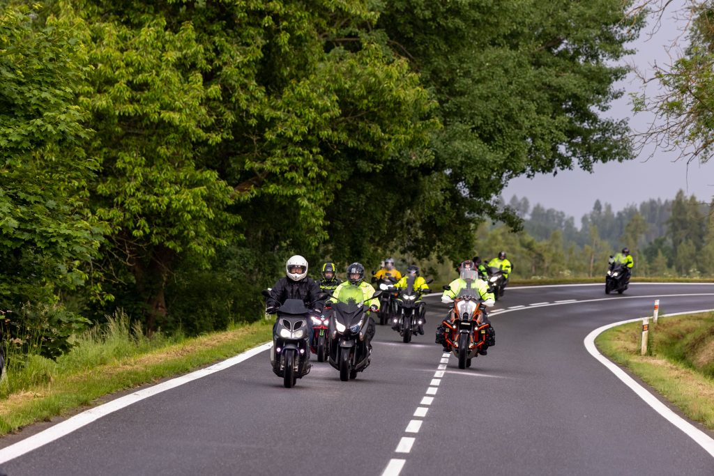 Skupina motocyklistů projíždí v koloně zatáčkou na silnici lemované stromy.