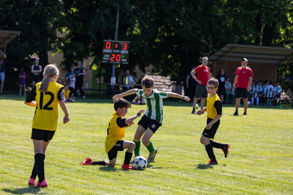 Mladá fotbalistka v zelenobílém dresu prochází mezi dvěma soupeřkami ve žlutém.