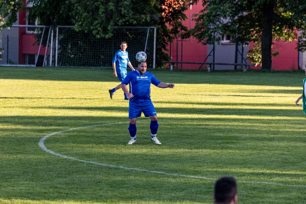 Fotbalista v modrém dresu zpracovává míč hrudí na sluncem osvětleném hřišti.