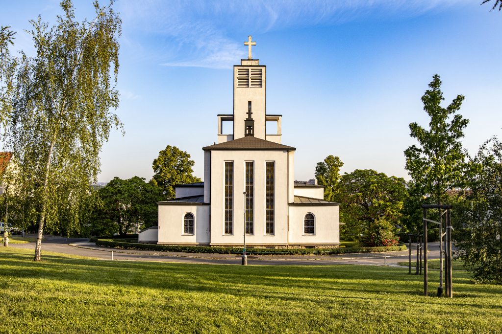 Moderní světlý kostel s hranolovou věží a křížem, obklopený trávníkem a stromy.
