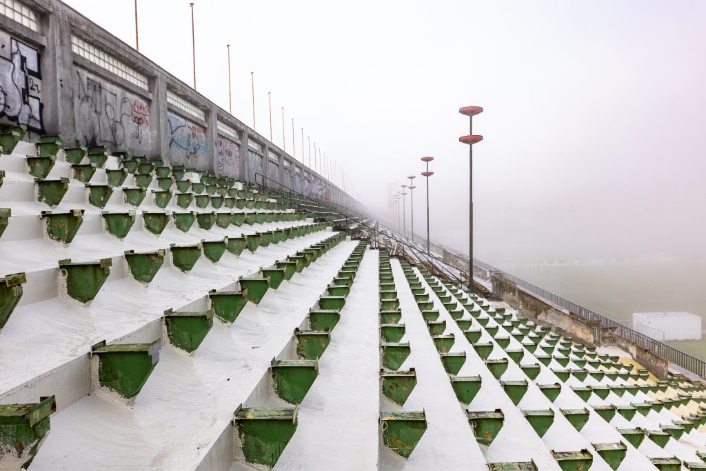 Prázdné řady zelenobílých sedadel venkovní tribuny mizící v husté mlze.
