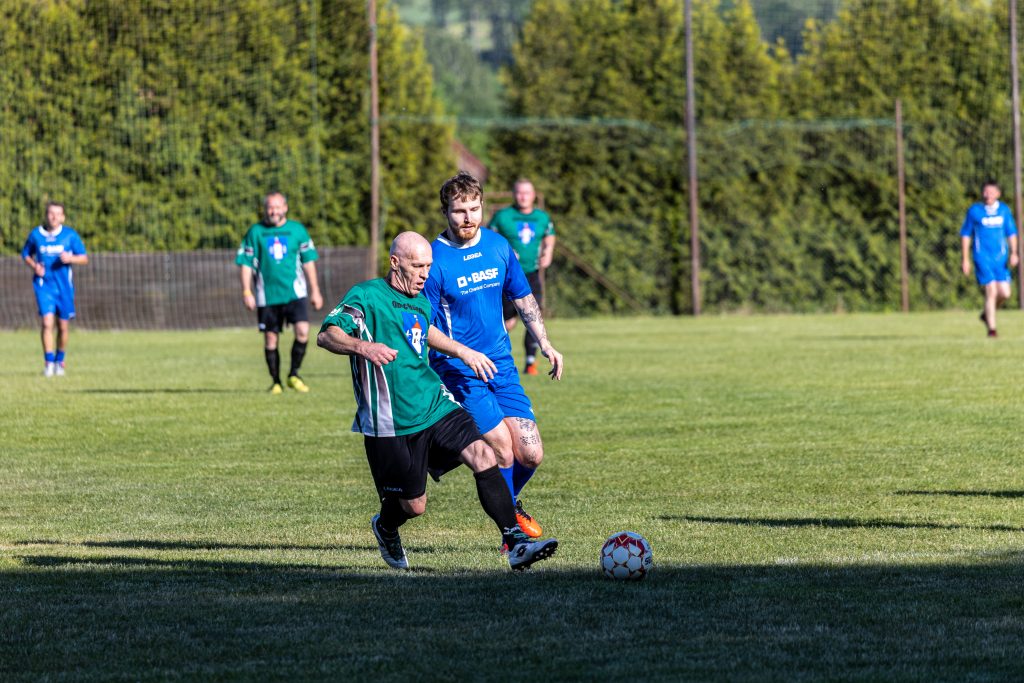 Fotbalista v zeleném kryje míč před soupeřem v modrém.