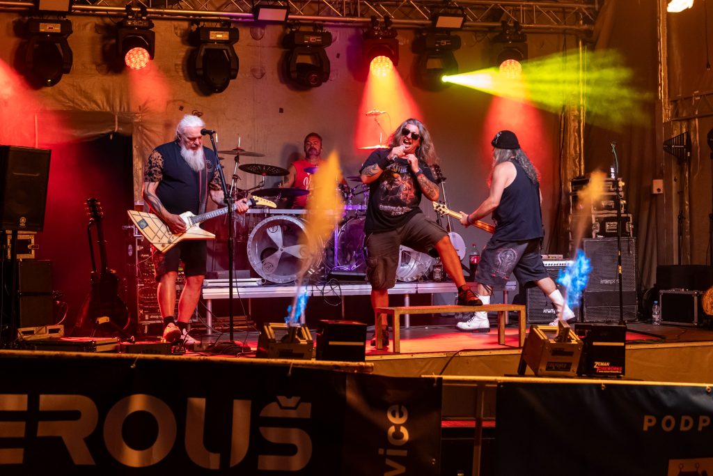 Čtyřčlenná rocková kapela hraje v širokém záběru pod červenými a žlutými světly, dva kytaristé se naklánějí k sobě.