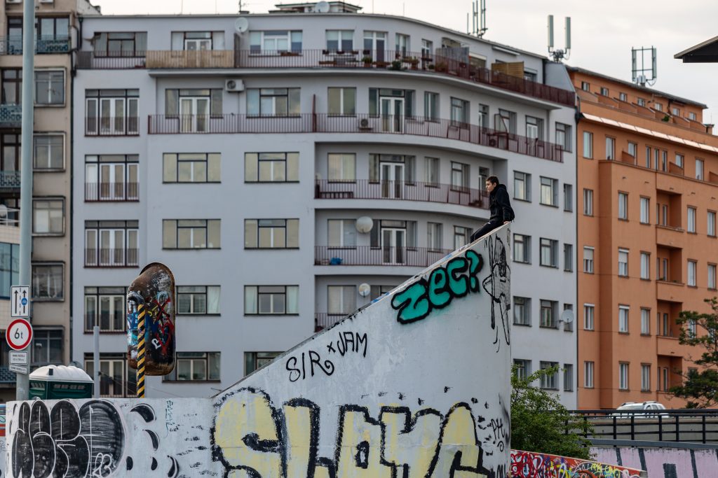Skateboardista ve výskoku nad betonovou rampou pokrytou graffiti, v pozadí městské domy.