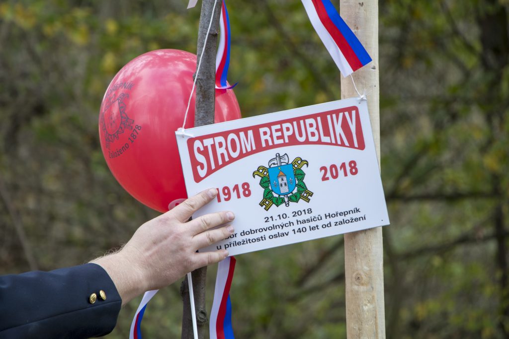 Ruka drží ceduli Strom republiky 1918–2018 u nově vysazené lípy ozdobené českou trikolorou.