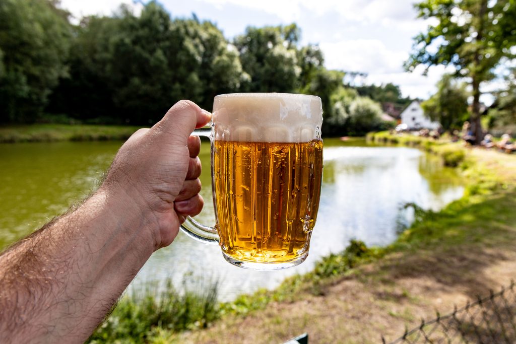 Ruka drží orosený půllitr piva nad břehem rybníka.