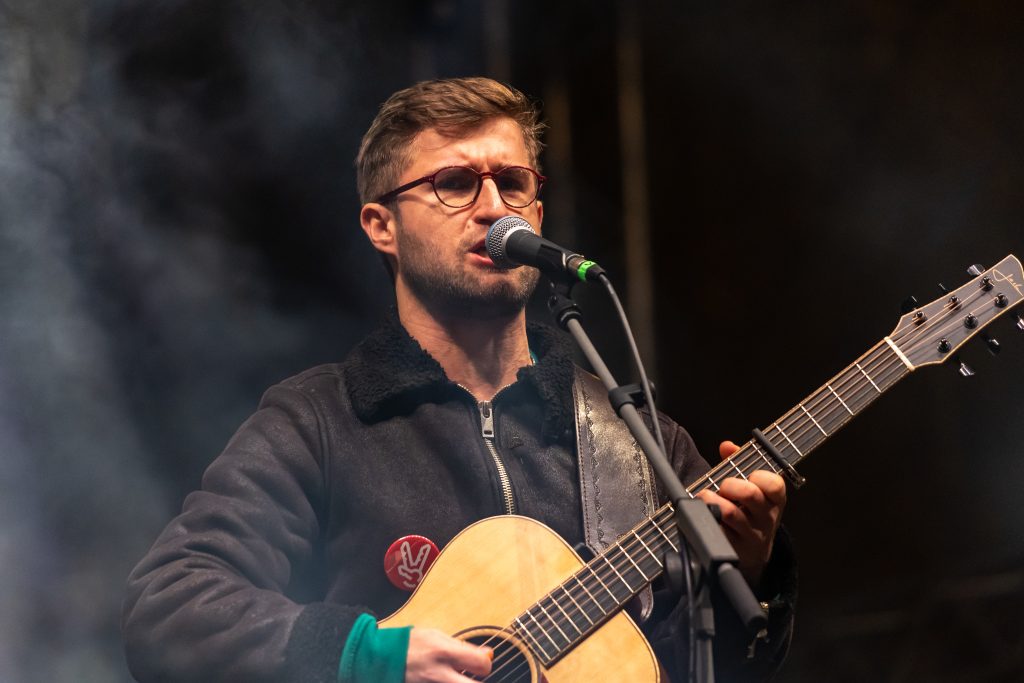 Muž s brýlemi zpívá do mikrofonu a hraje na akustickou kytaru proti tmavému zakouřenému pozadí.