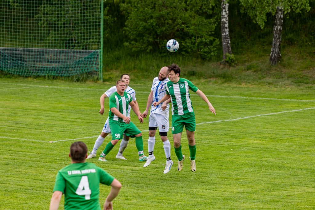 Skupina fotbalistů skáče za vysokým míčem před brankou.