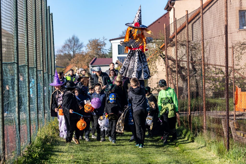 Průvod dětí a dospělých v čarodějnických a strašidelných kostýmech na travnaté cestě.