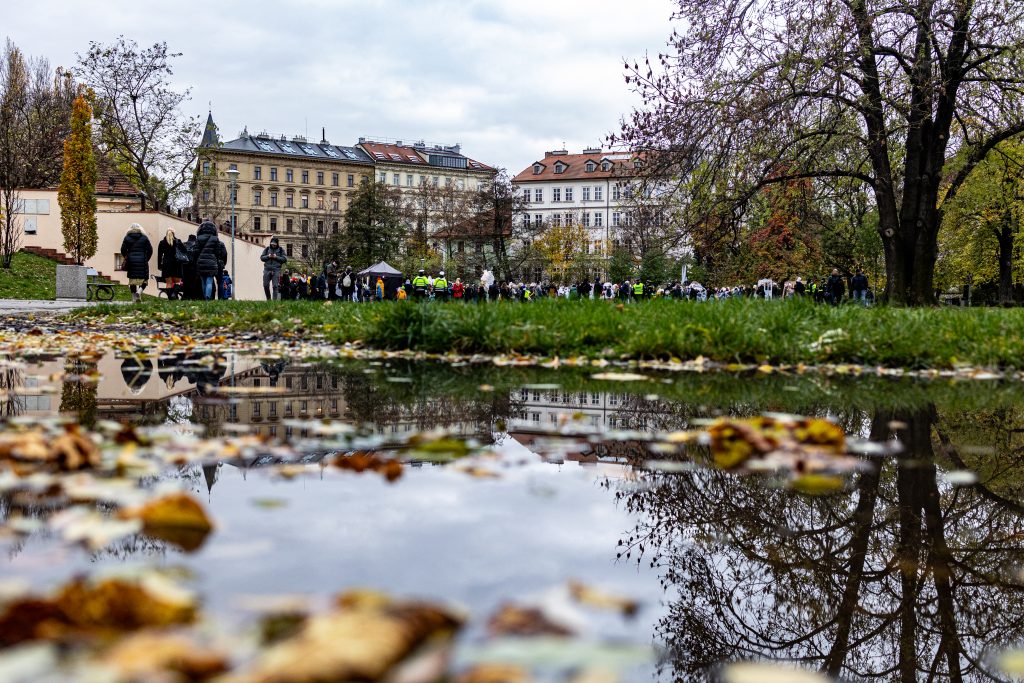 Podzimní park s louží v popředí, ve které se odrážejí stromy, a zástupem lidí v pozadí.