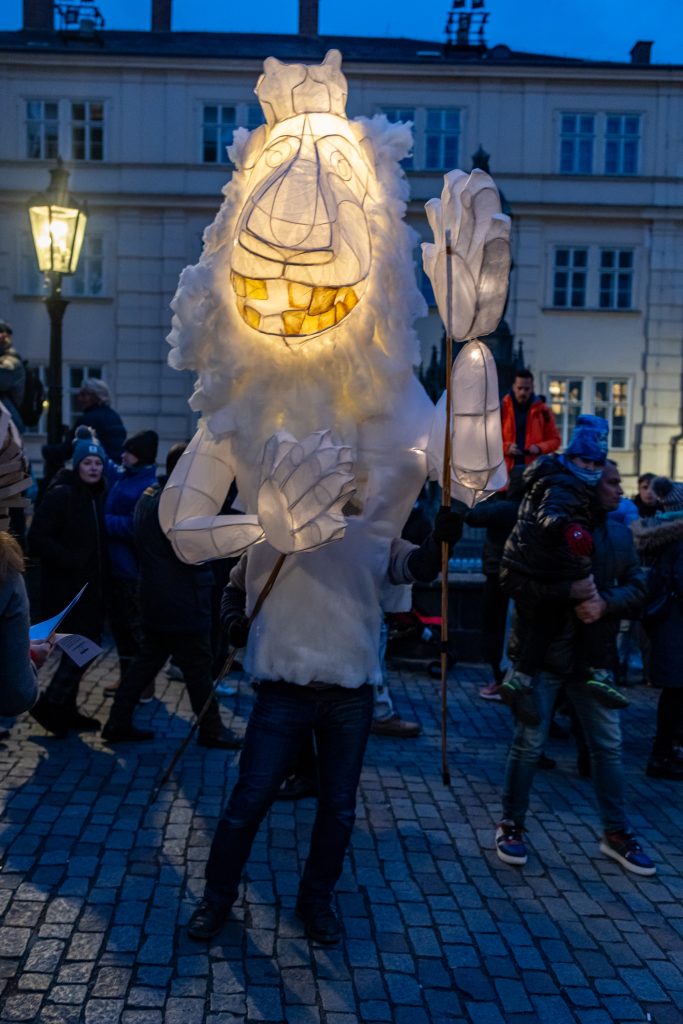 Účastník průvodu nese velkou bílou svítící masku koně mezi lidmi na Karlově mostě.