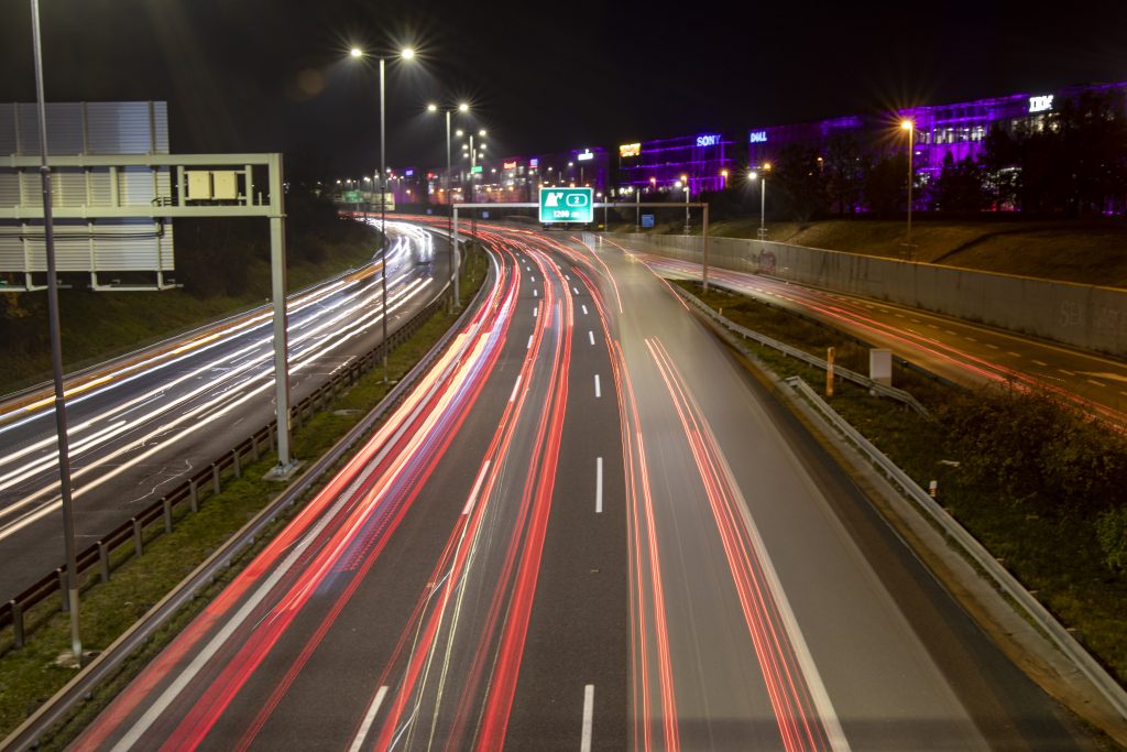 Světelné stopy aut na noční víceproudé komunikaci, nad ní osvětlené obchodní budovy.
