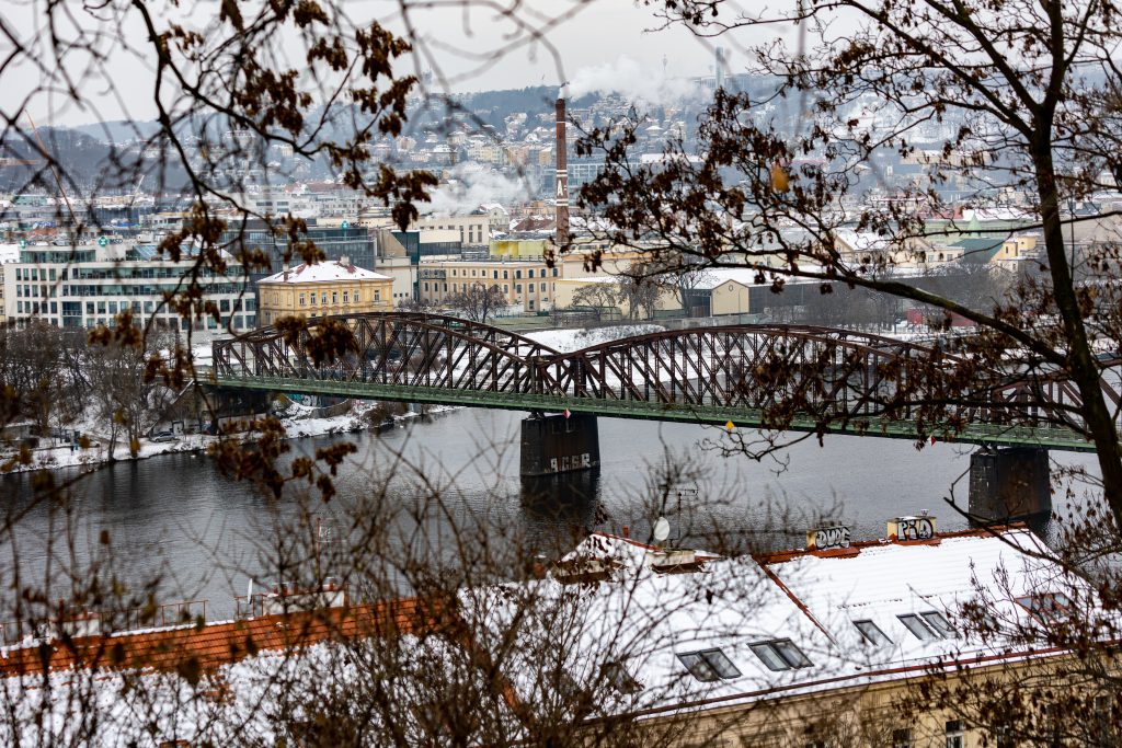 Železniční most přes Vltavu a pražské nábřeží pozorované skrz větve ze svahu.