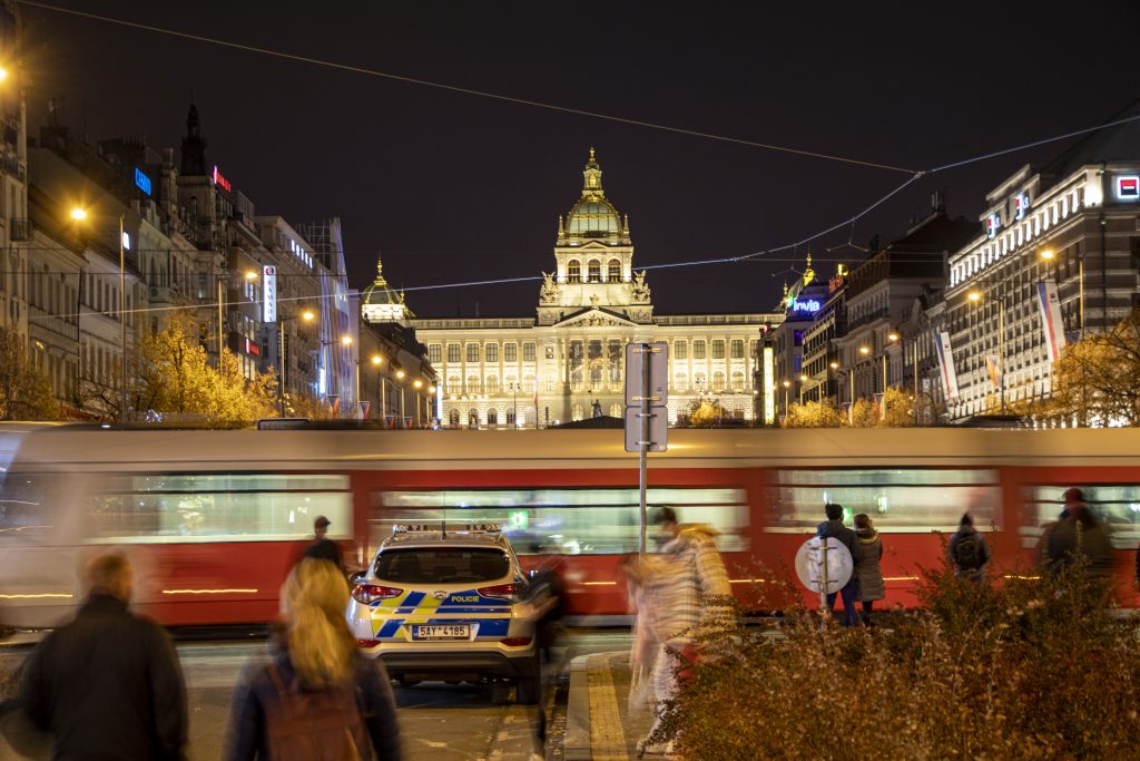 Rozmazaná červenobílá tramvaj projíždí před osvětleným Národním muzeem a policejním vozem.